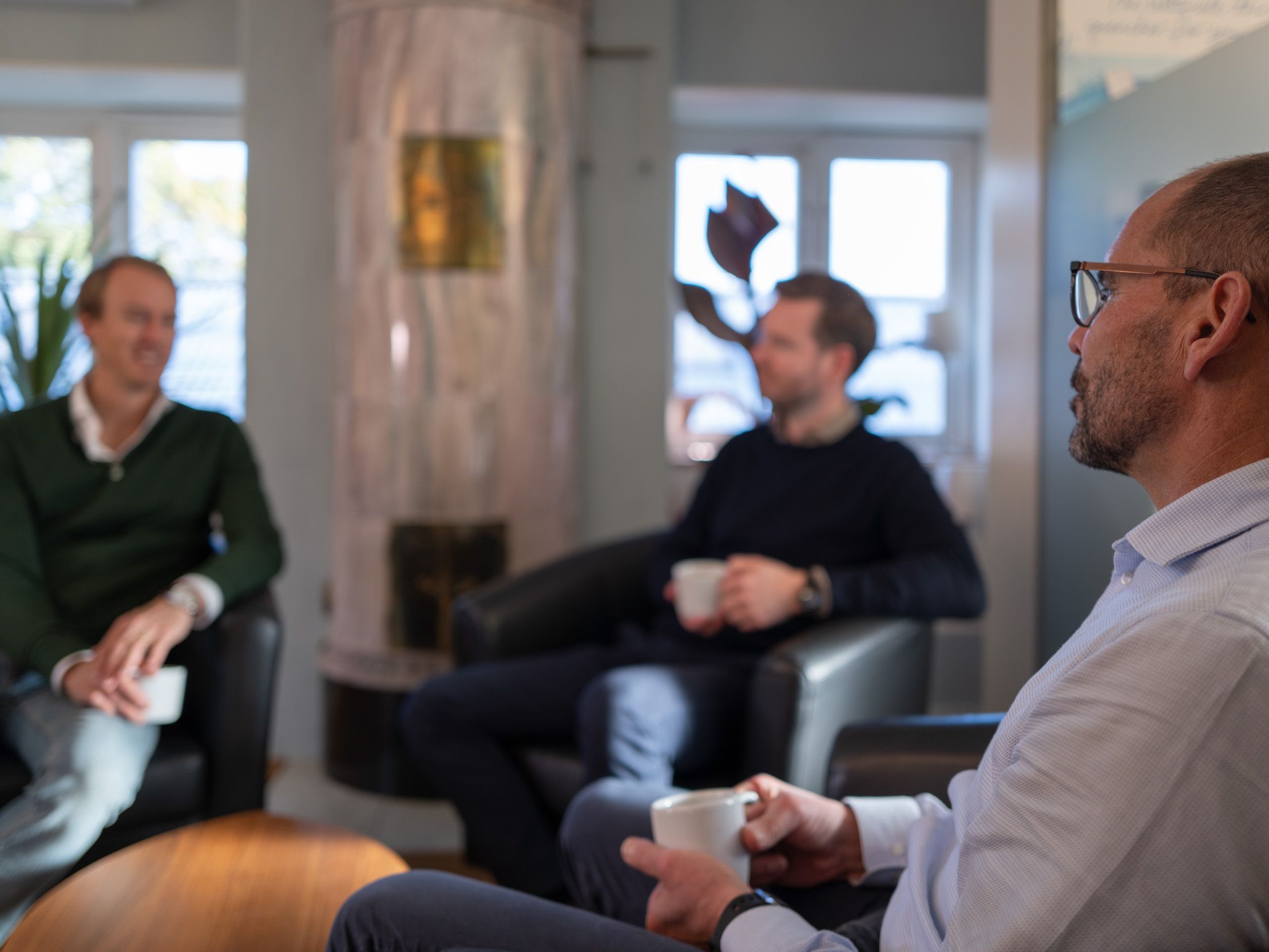 Tre personer diskuterar anställning. De sitter i fåtöljer i ett mötesrum, framför en kakelspis. De håller varsin kaffekopp och de lyssnar intresserat på varandra.