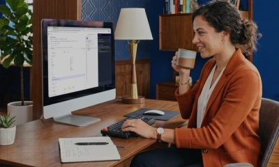 A woman working at a modern wooden desk with a large computer monitor while drinking from a mug.