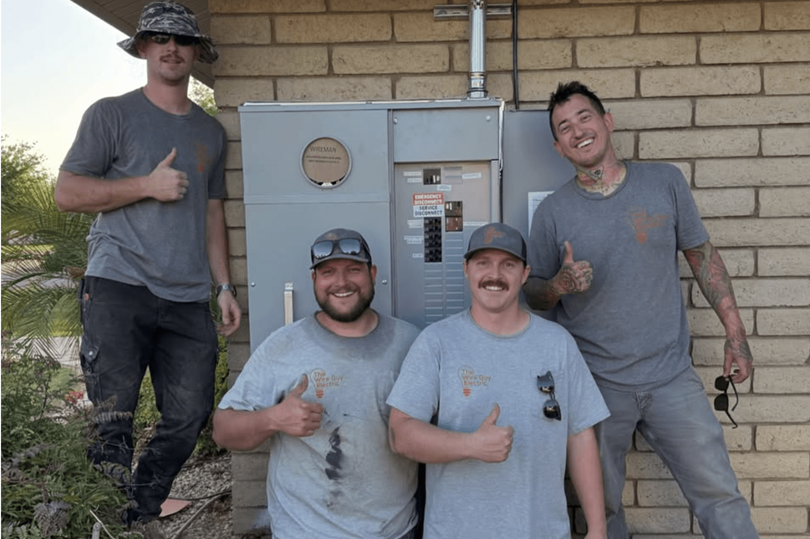 Four Wire Guy Electric electricians in company T-shirts giving a thumbs-up beside a new meter and panel on a block wall