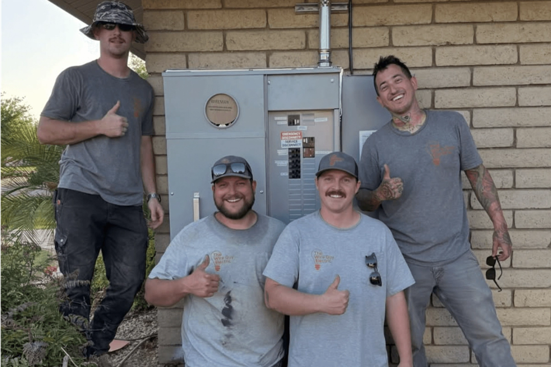 Four Wire Guy Electric electricians in company T-shirts giving a thumbs-up beside a new meter and panel on a block wall