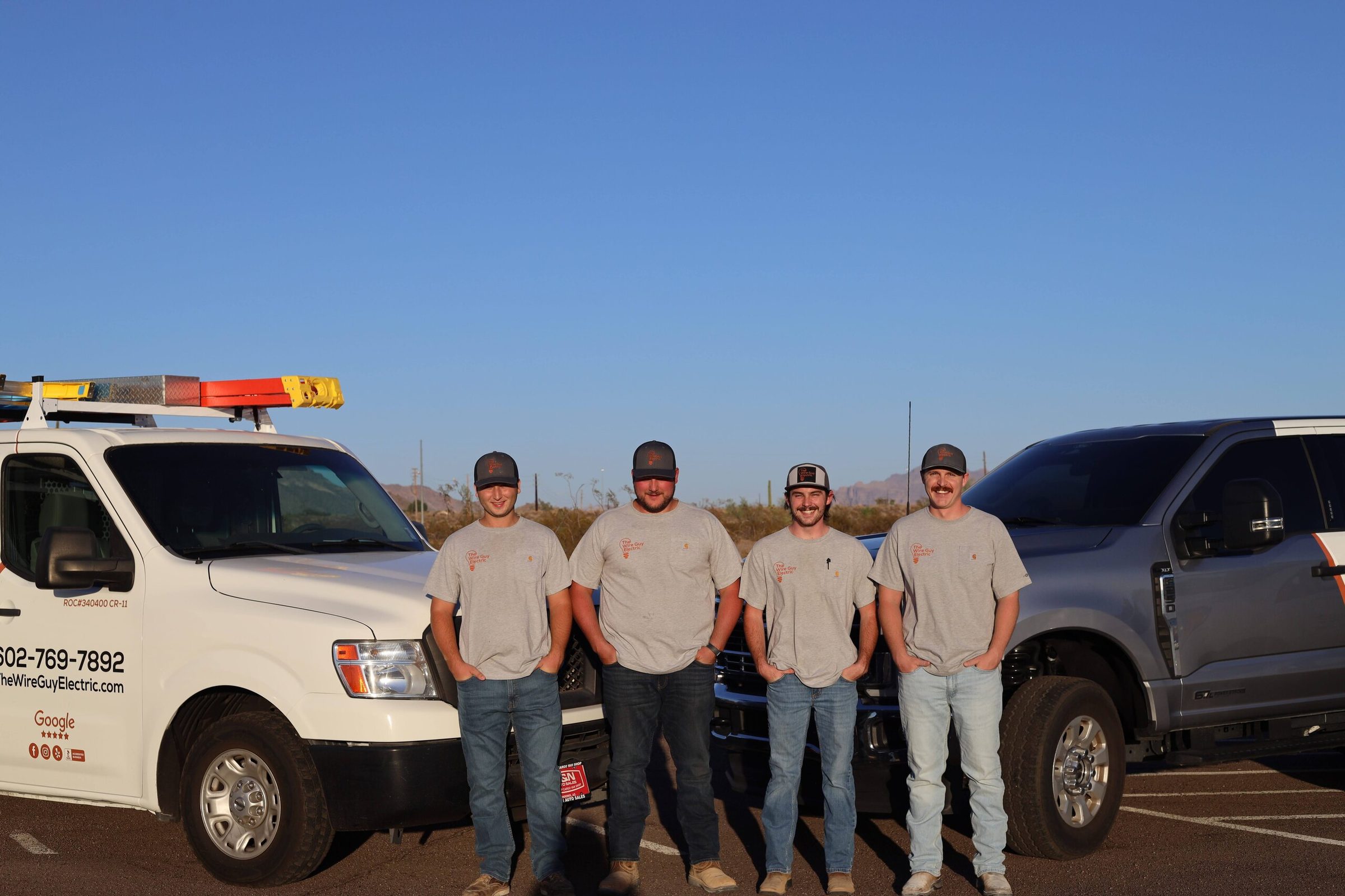 Four Wire Guy Electric technicians standing between a branded work van and pickup in the desert