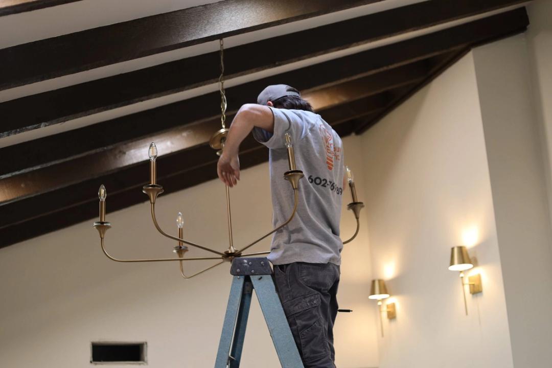 Electrician on a ladder installing a brass chandelier with lit wall sconces nearby