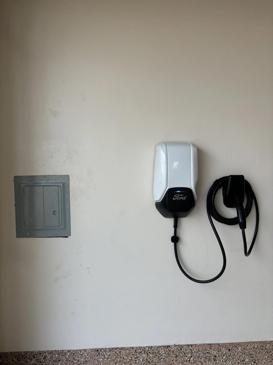 Ford wall-mounted EV charger with coiled cable next to a subpanel in a garage