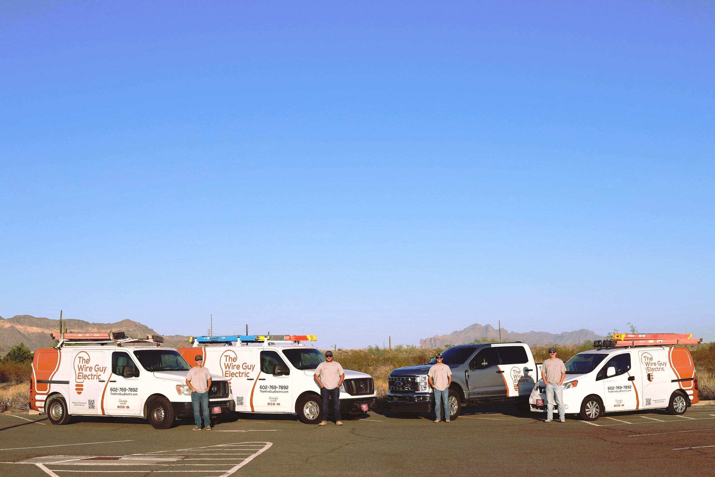 Three Wire Guy Electric vans and a company pickup lined up in the desert, with four electricians and the mountains behind them