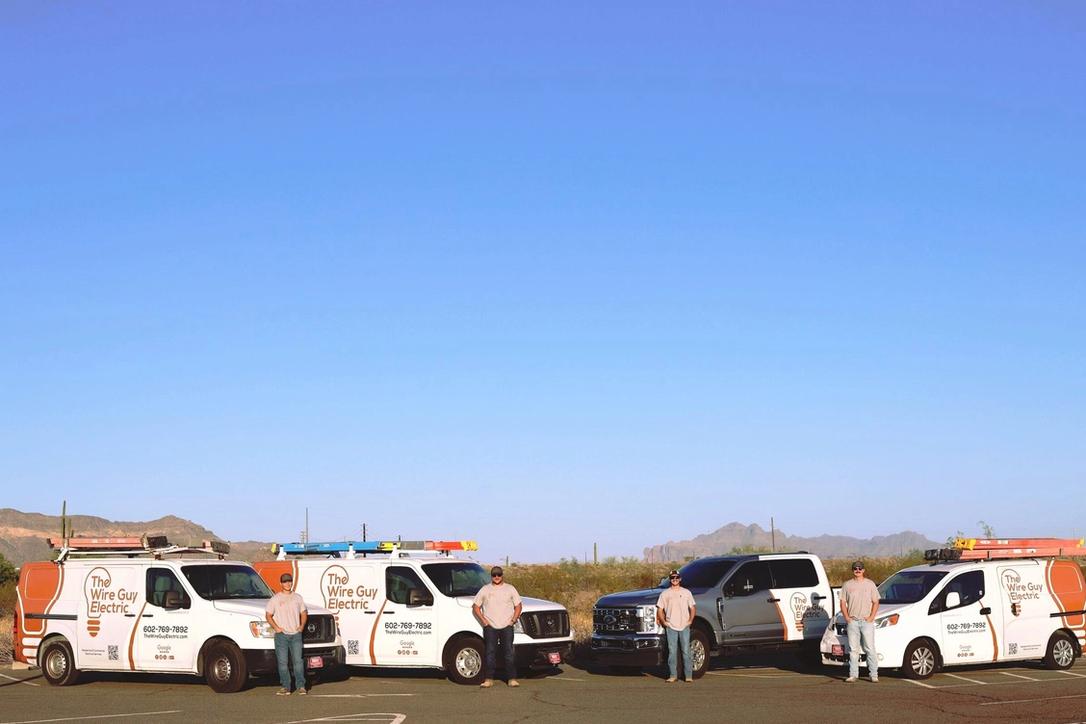Three Wire Guy Electric vans and a company pickup lined up in the desert, with four electricians and the mountains behind them
