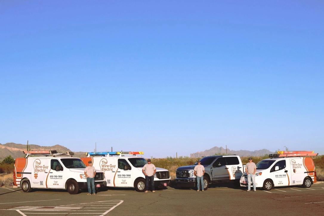 Three Wire Guy Electric vans and a company pickup lined up in the desert, with four electricians and the mountains behind them