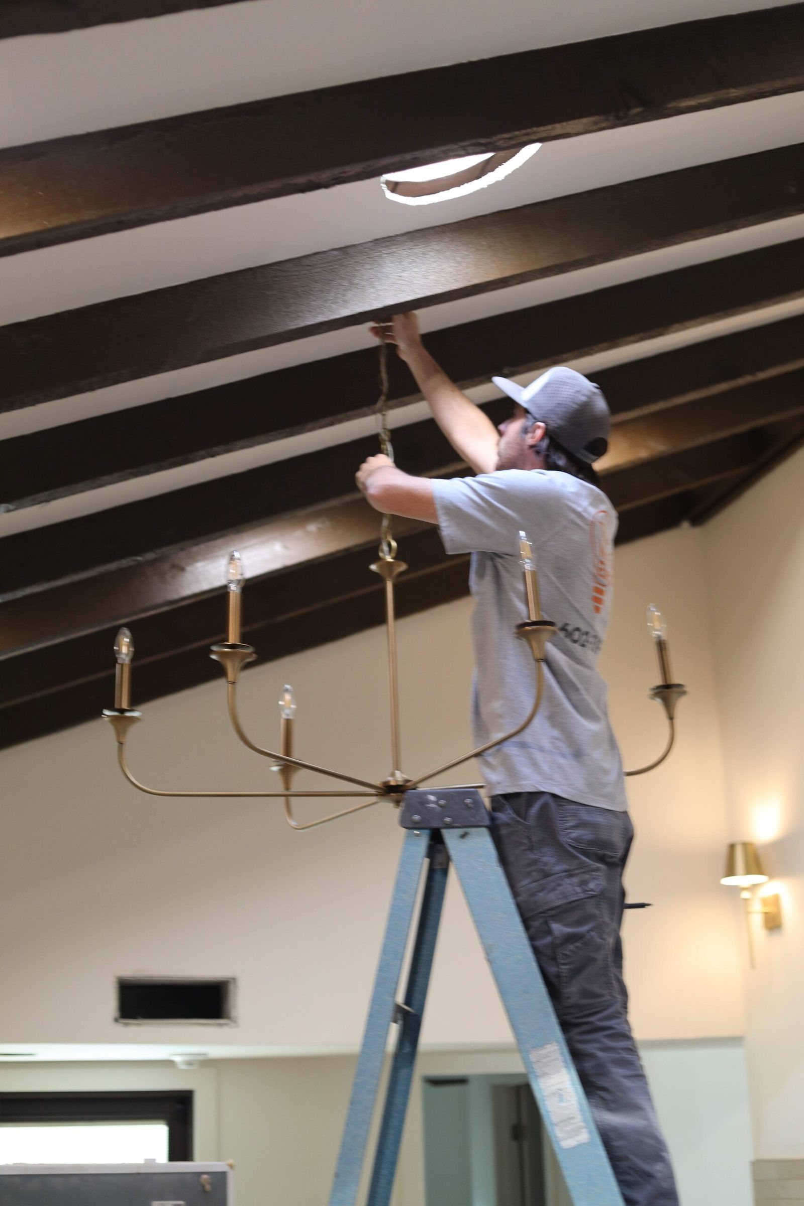 Electrician on a ladder hanging a brass candle-style chandelier from a dark beamed ceiling