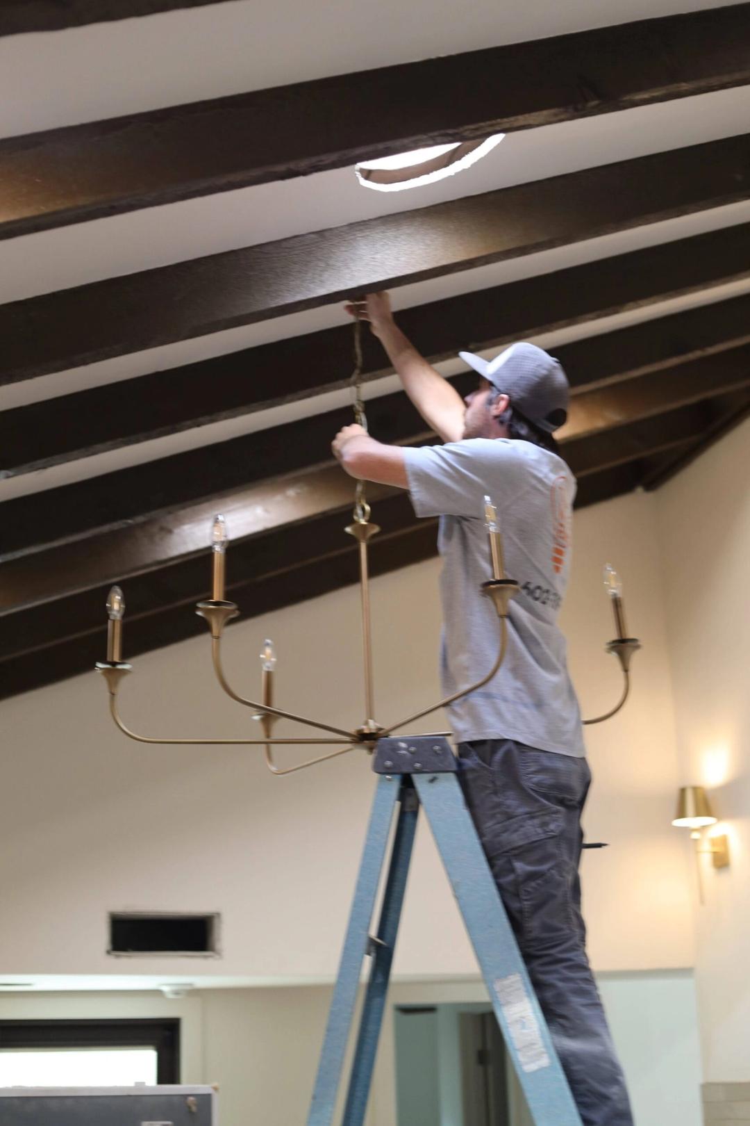 Electrician on a ladder hanging a brass candle-style chandelier from a dark beamed ceiling