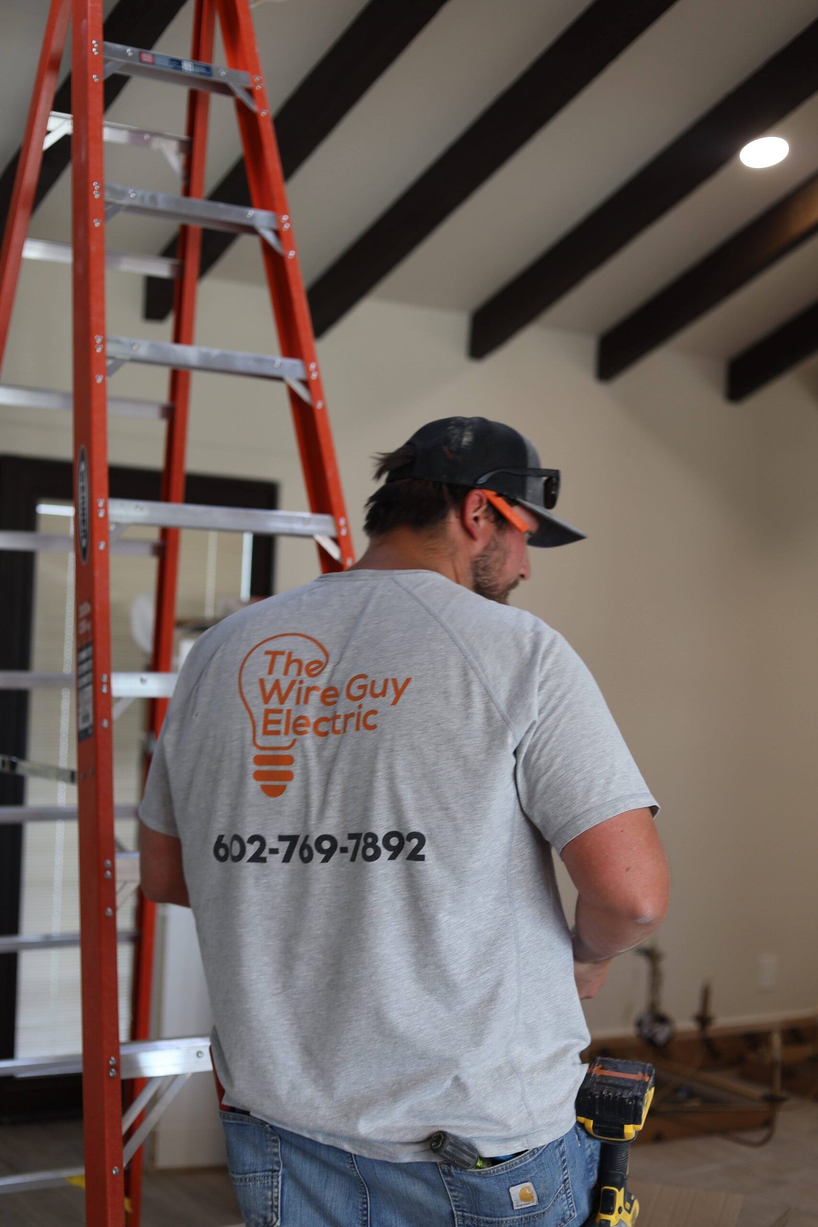Electrician seen from behind in a Wire Guy Electric shirt with phone number beside a tall orange ladder