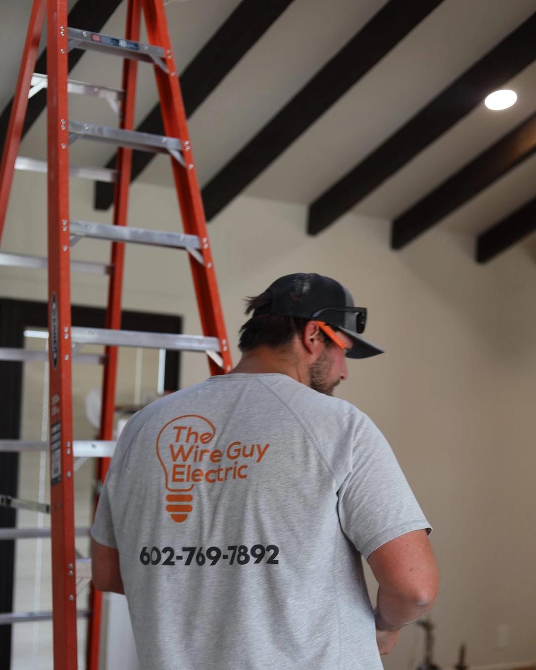 Electrician seen from behind in a Wire Guy Electric shirt with phone number beside a tall orange ladder
