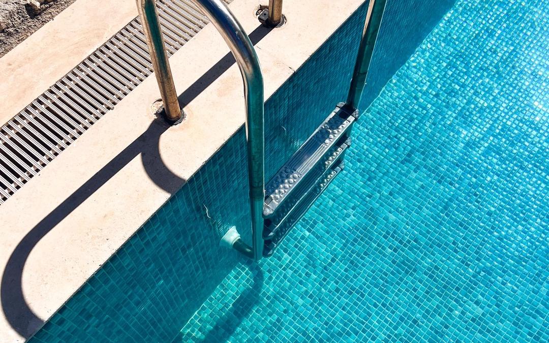 Steel ladder rails leading into a blue-tiled swimming pool, with a drain grate along its edge