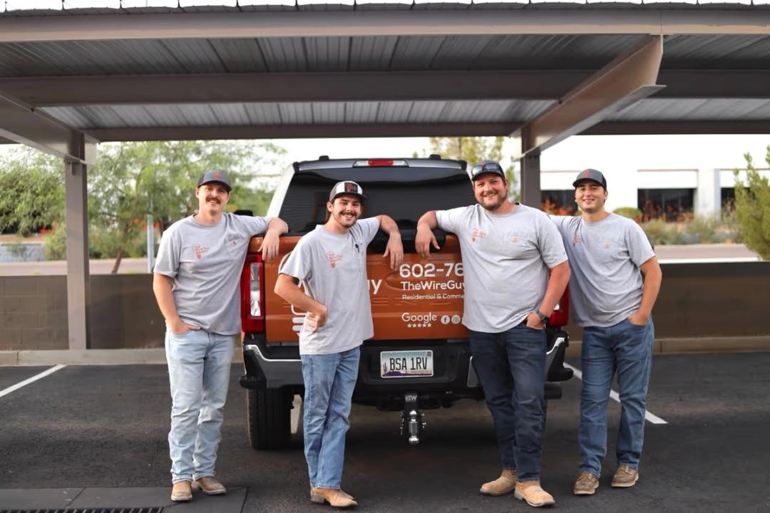 Four Wire Guy Electric technicians leaning on the tailgate of the company's copper-wrapped pickup under a carport