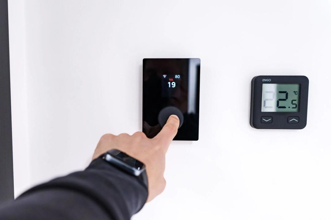 A hand pressing the dial of a black smart thermostat beside a second digital thermostat