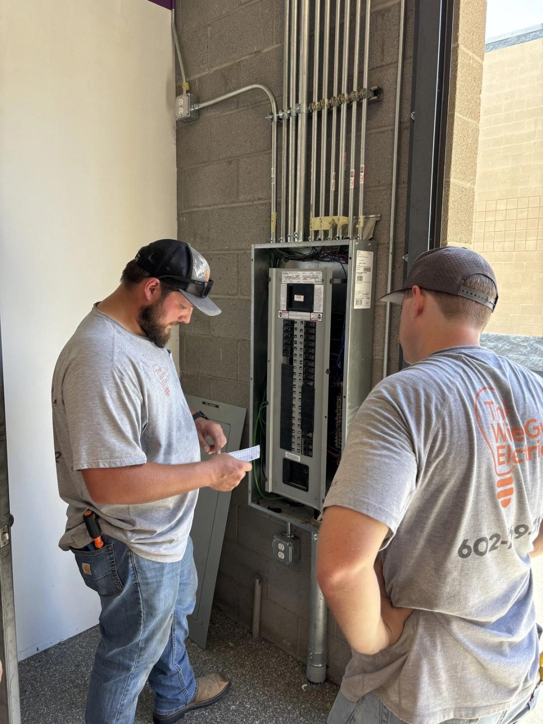 Two electricians in company shirts checking a commercial panel with conduit runs