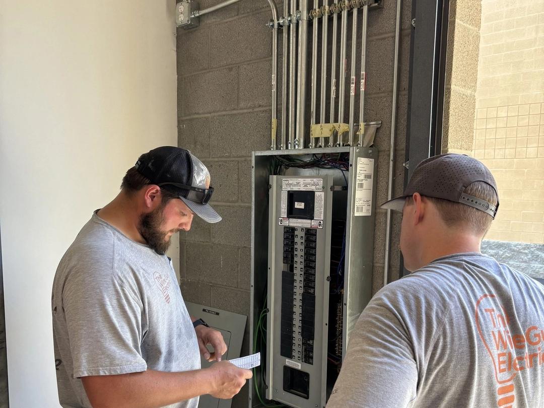 Two electricians in company shirts checking a commercial panel with conduit runs