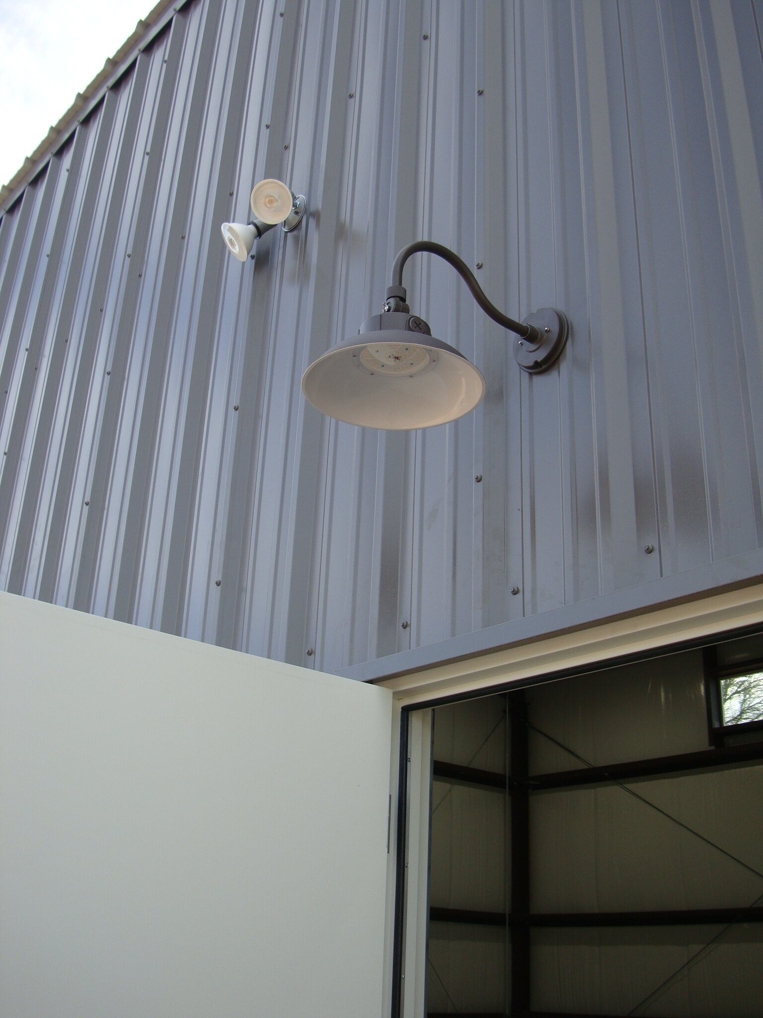Black gooseneck barn light and floodlight on a gray metal building above a door