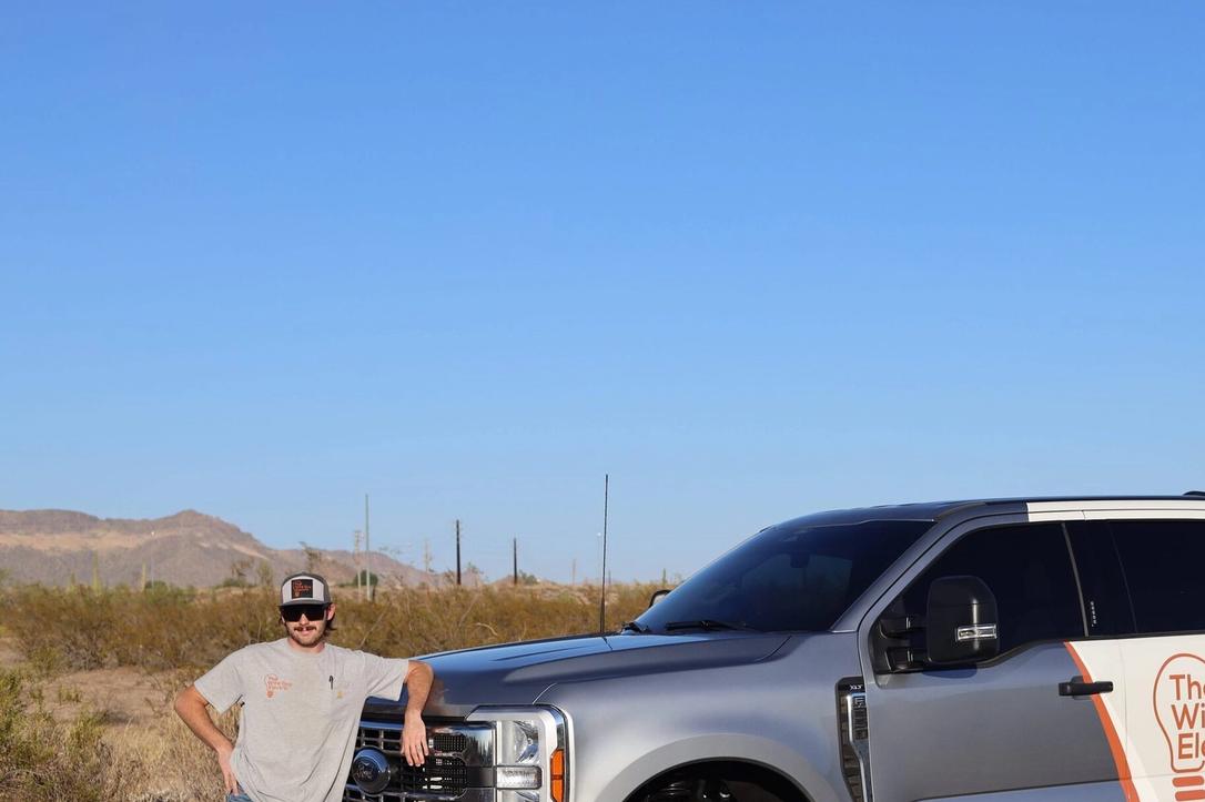 Wire Guy Electric technician in sunglasses leaning on a branded silver and white pickup in the desert