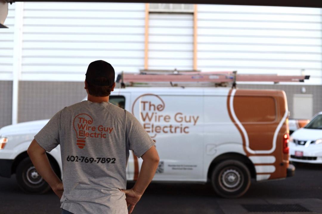 A Wire Guy electrician, seen from behind in a company shirt with the phone number on the back, looking at a company van