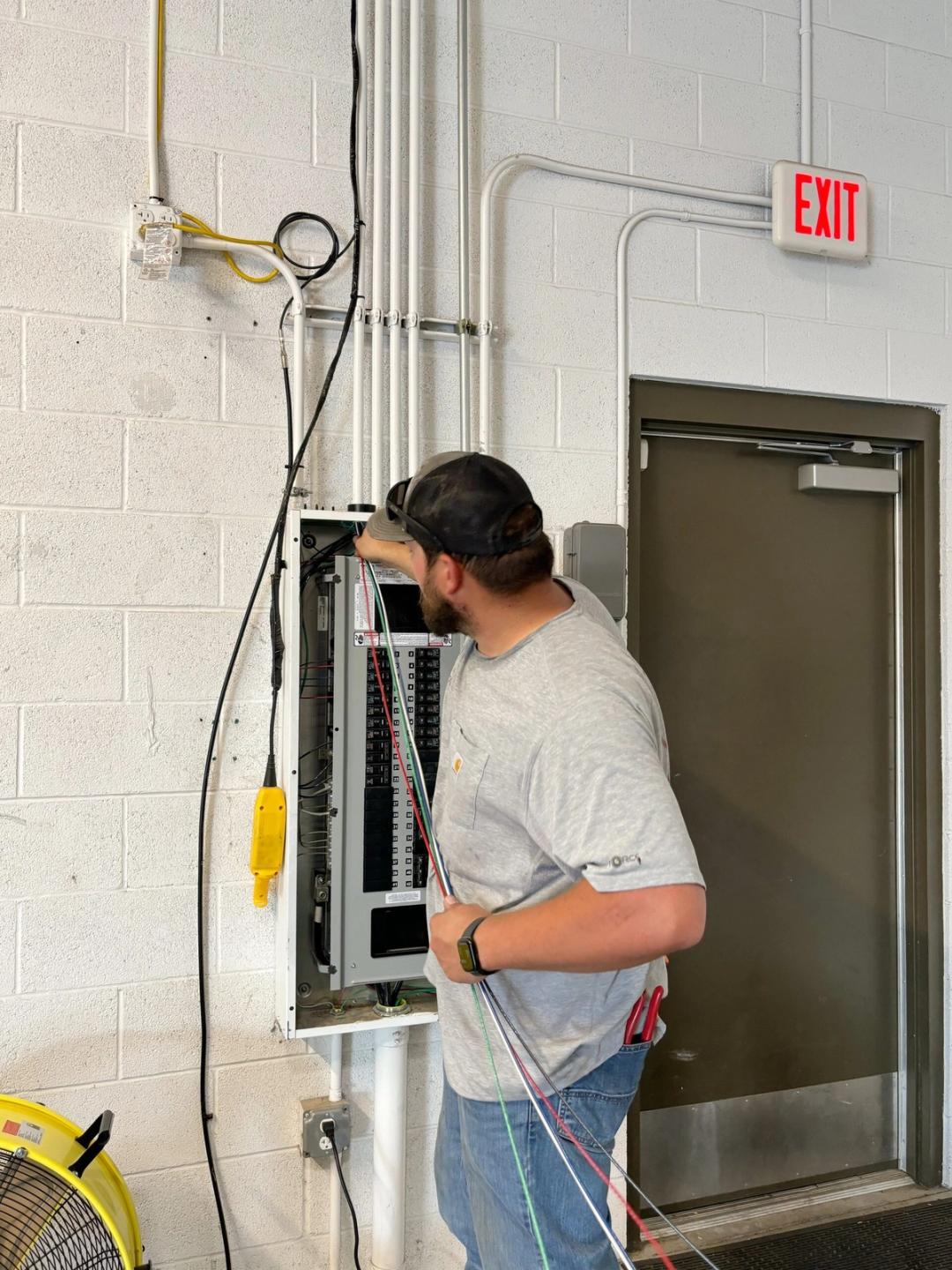 Electrician wiring a commercial panel on a block wall beside an exit sign