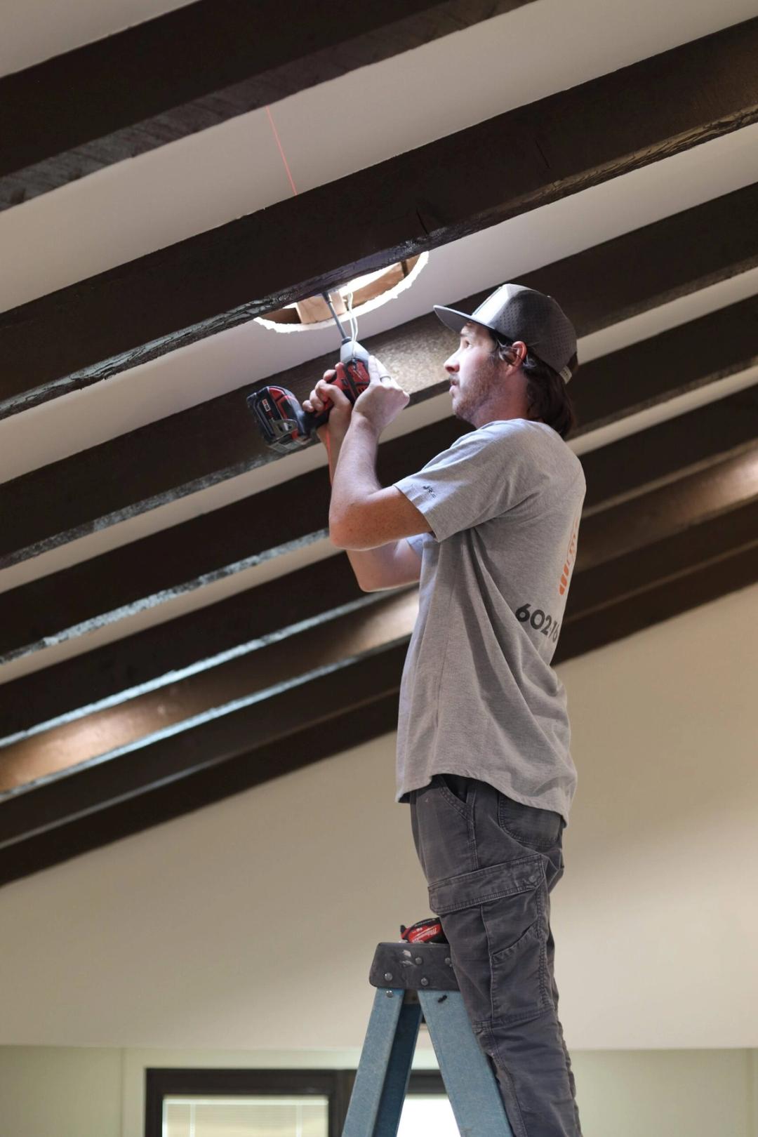 Electrician drilling into a dark beamed ceiling at a recessed light opening