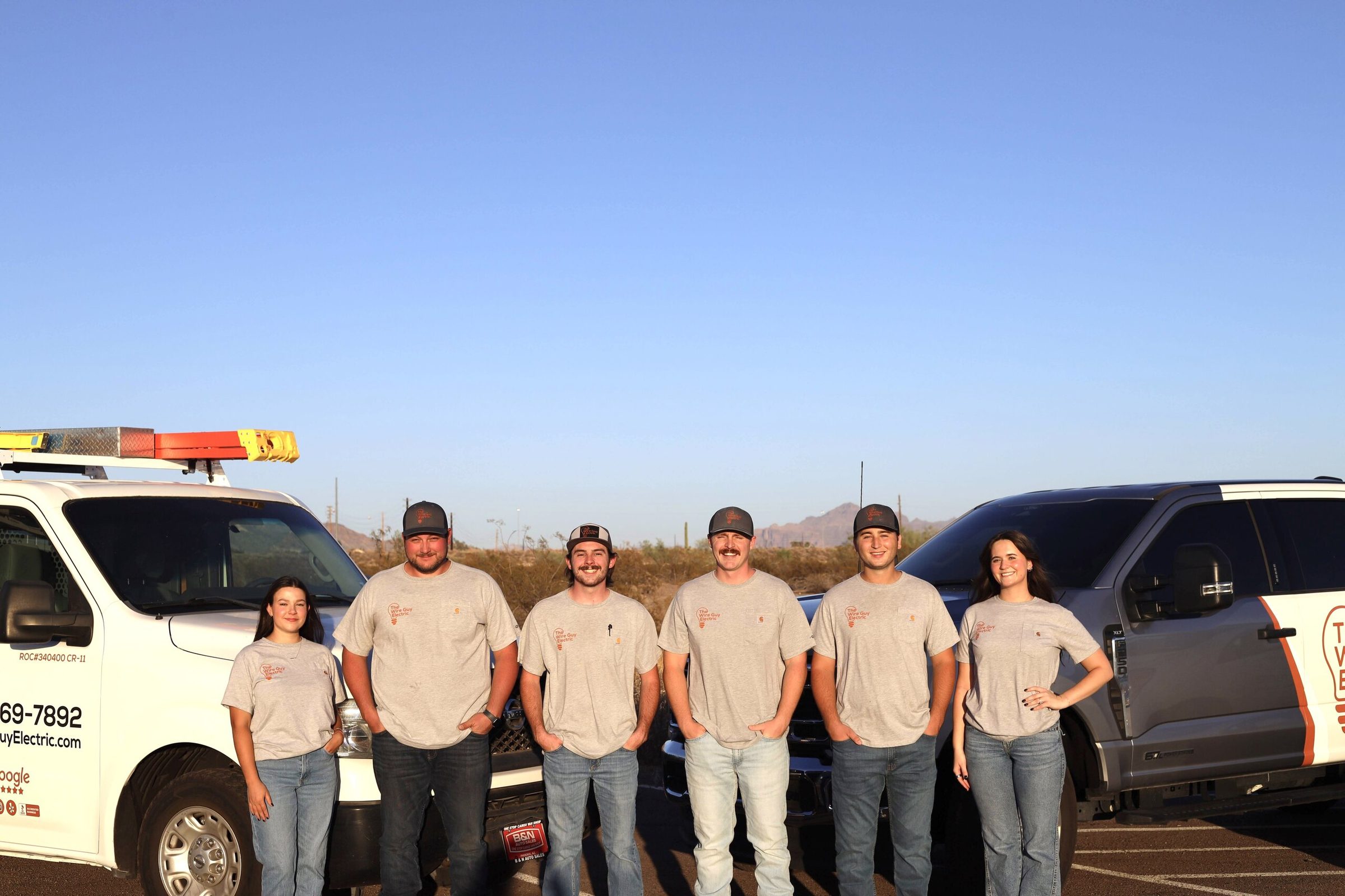 Six Wire Guy Electric team members in company shirts beside vans under a clear evening sky