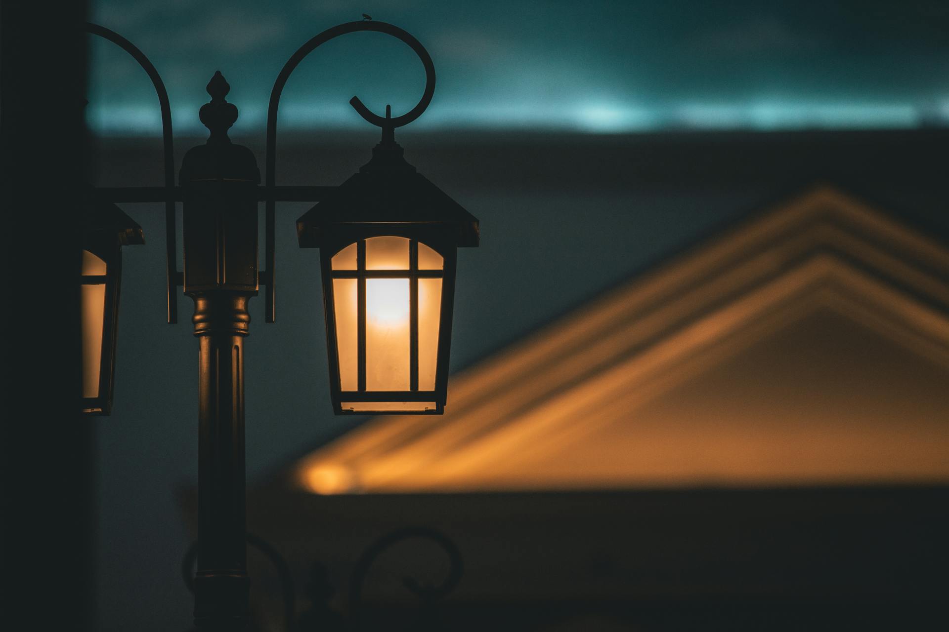A lit lantern-style lamp post at dusk, with a roofline glowing behind it