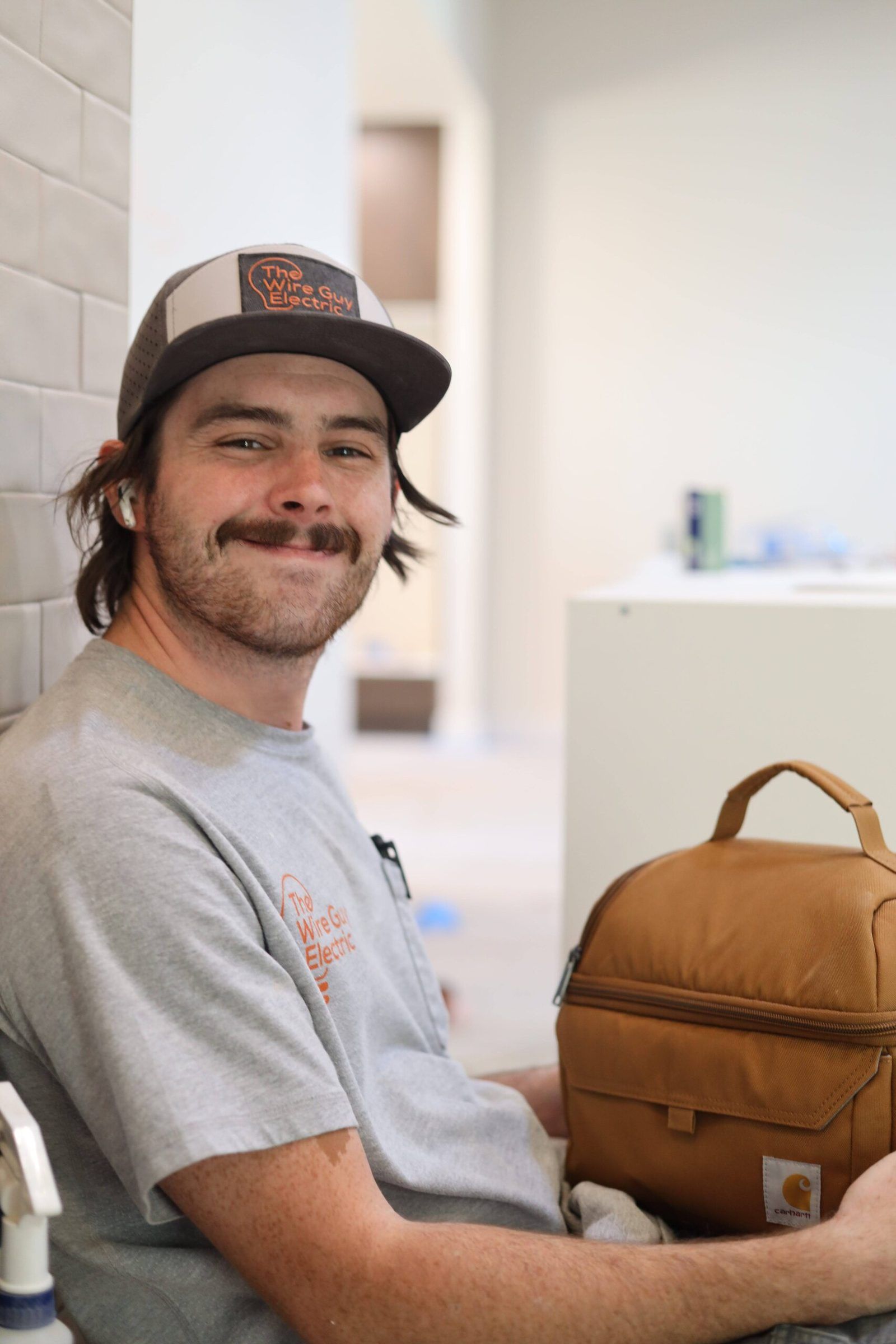 Smiling technician in a Wire Guy Electric cap and shirt sitting with a tan lunch bag