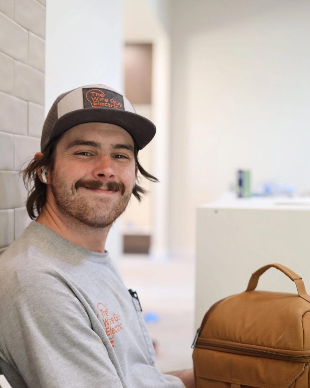 Smiling technician in a Wire Guy Electric cap and shirt sitting with a tan lunch bag