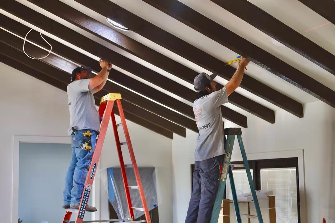 Two Wire Guy Electric electricians on ladders installing lighting in a room with a vaulted, dark-beamed ceiling