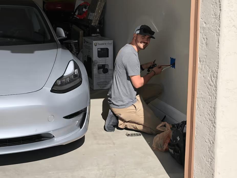 A Wire Guy Electric electrician kneeling beside a Tesla Model 3 in a home garage, wiring a box on the wall
