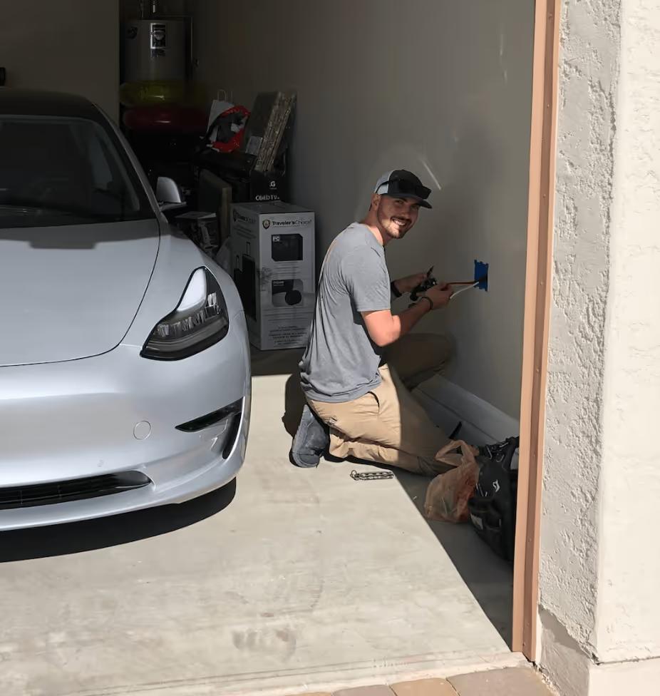 Wire Guy Electric electrician kneeling beside a Tesla Model 3 in a home garage, wiring a box on the wall