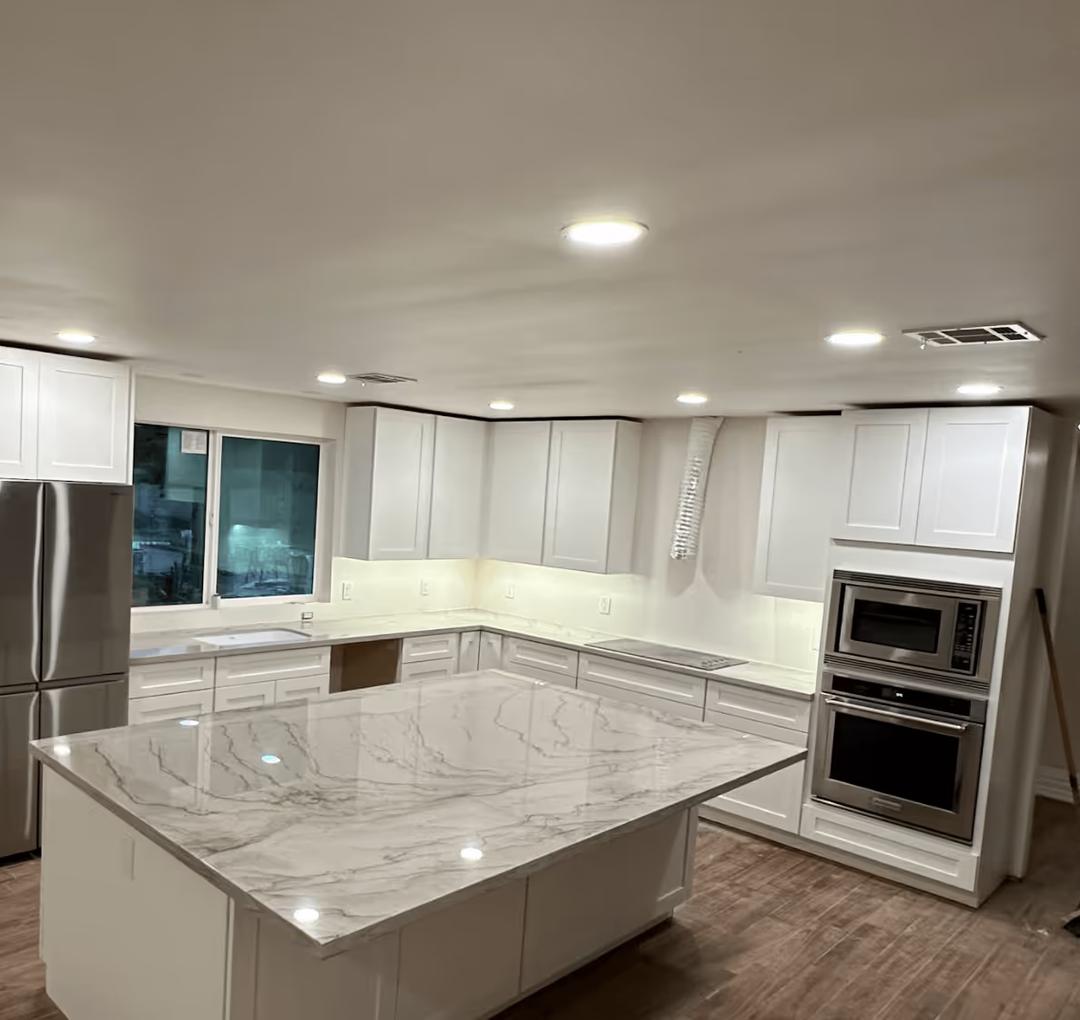 Remodeled kitchen at night lit by recessed ceiling lights, with white shaker cabinets and a marble-look island