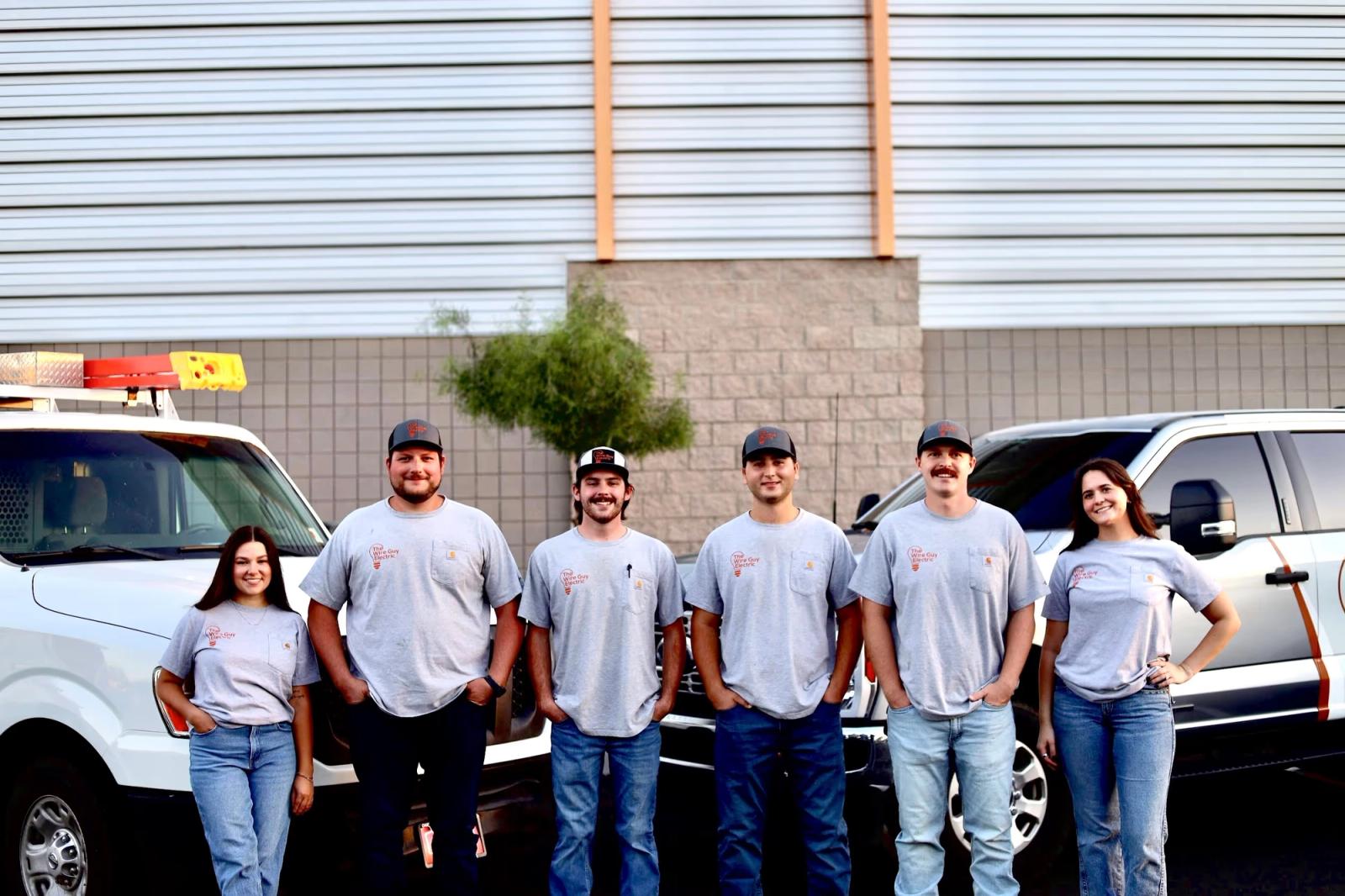 The Wire Guy Electric crew of six in matching gray company T-shirts, lined up in front of two white work trucks at the shop
