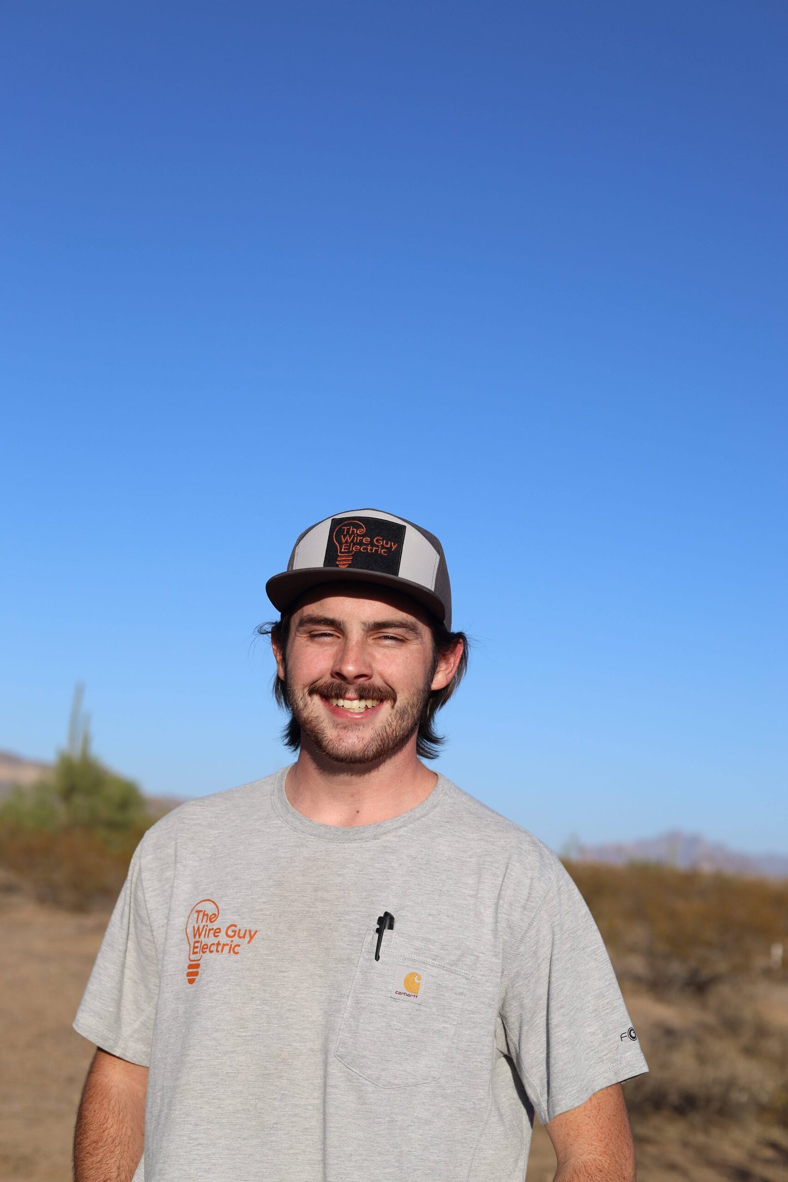 Grinning technician in a Wire Guy Electric cap and shirt against a blue desert sky