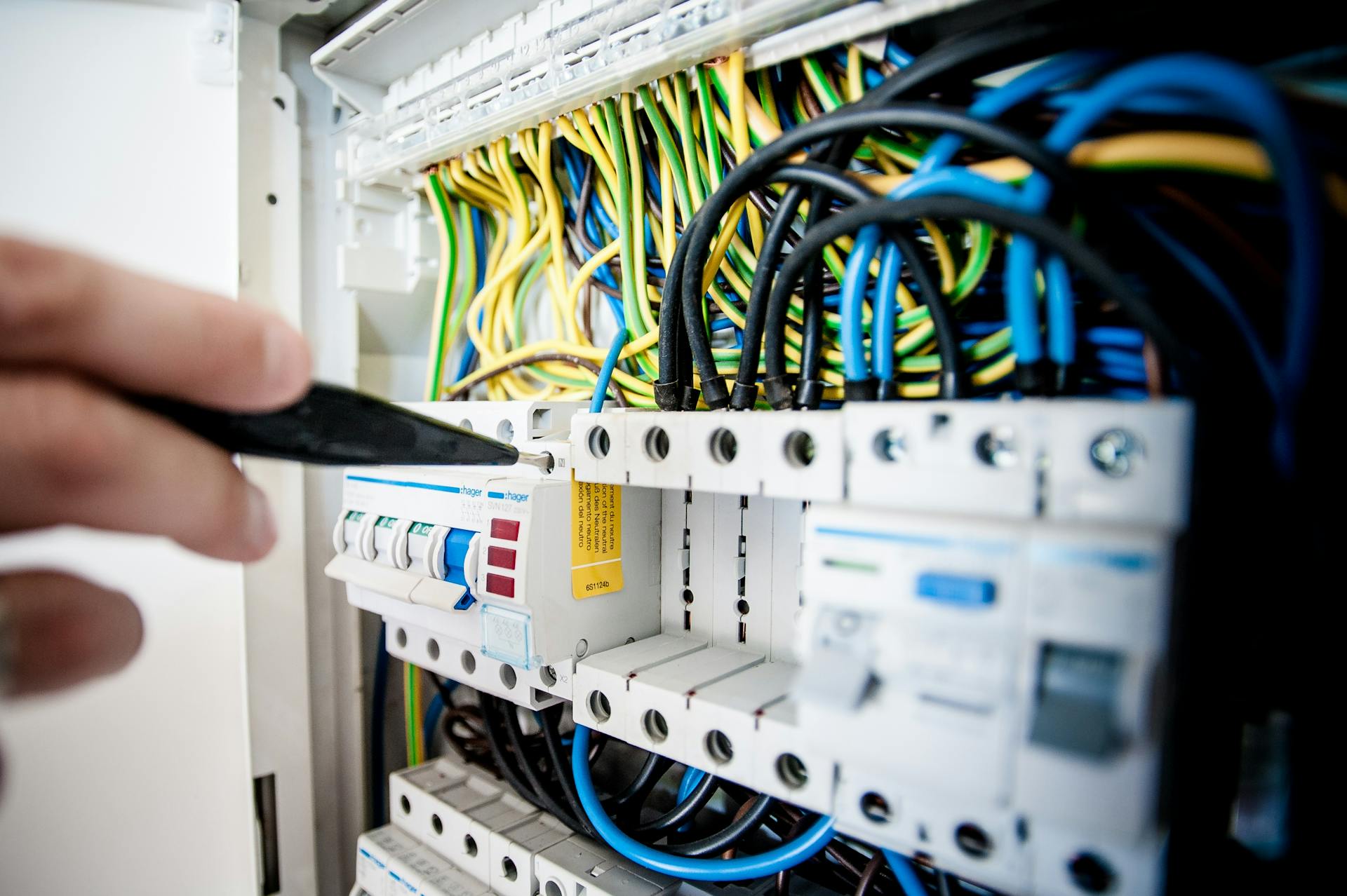 A hand holding a screwdriver to a breaker in an open distribution board full of blue, yellow and black wires