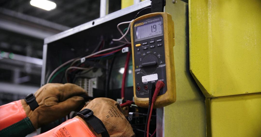 Gloved hands testing the wiring of a control panel with a yellow digital multimeter