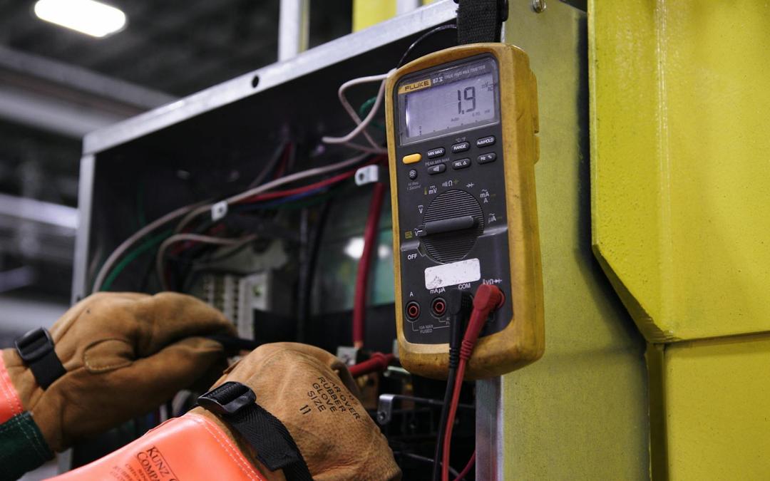 Gloved hands testing the wiring of a control panel with a yellow digital multimeter