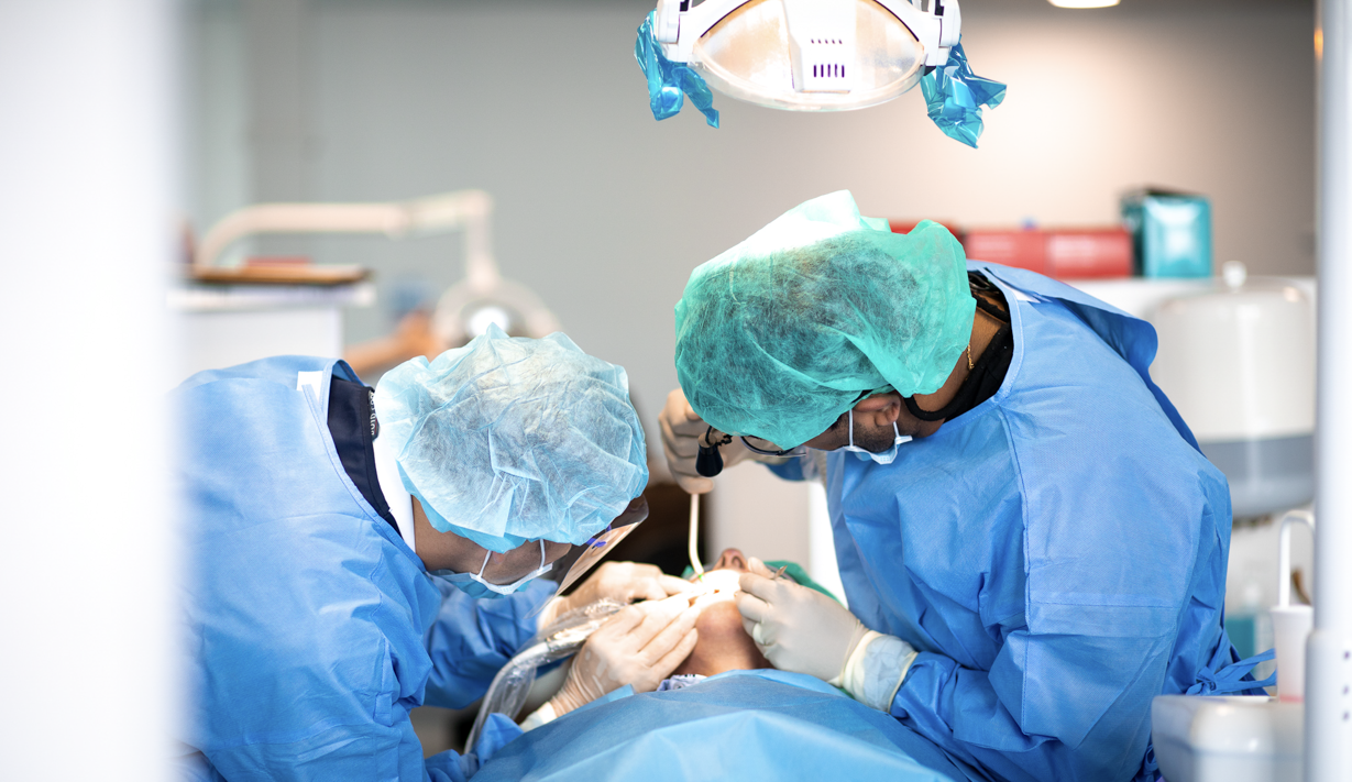 Two dental professionals in full surgical attire perform a dental procedure on a patient under an overhead surgical light in a modern dental operatory.