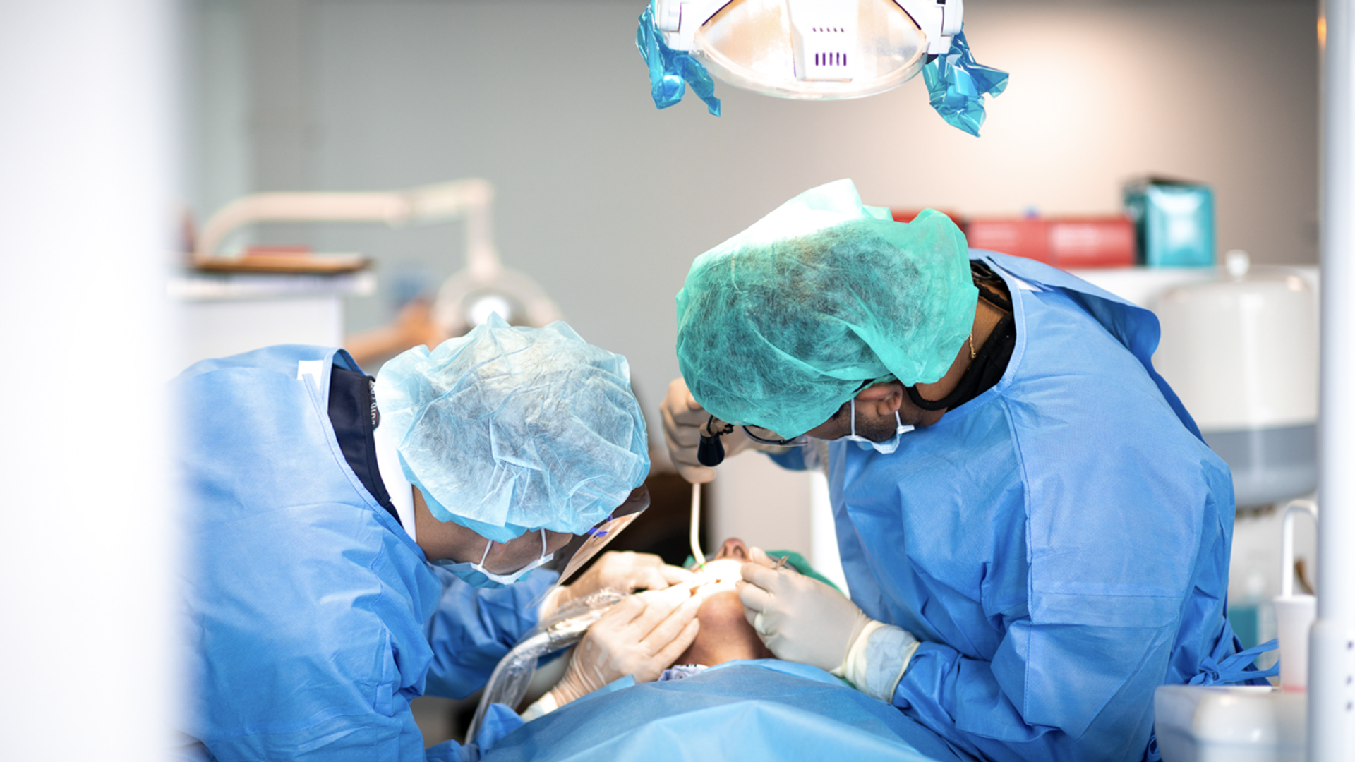 Two dental professionals in full surgical attire perform a dental procedure on a patient under an overhead surgical light in a modern dental operatory.