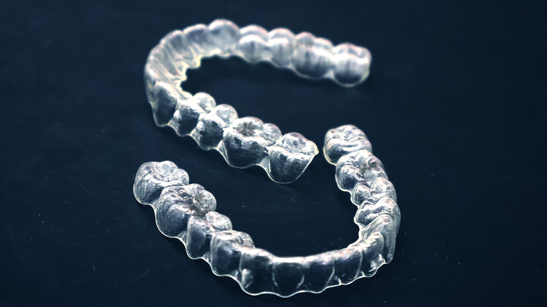 Two clear orthodontic aligner trays on a dark background showing tooth-form detail