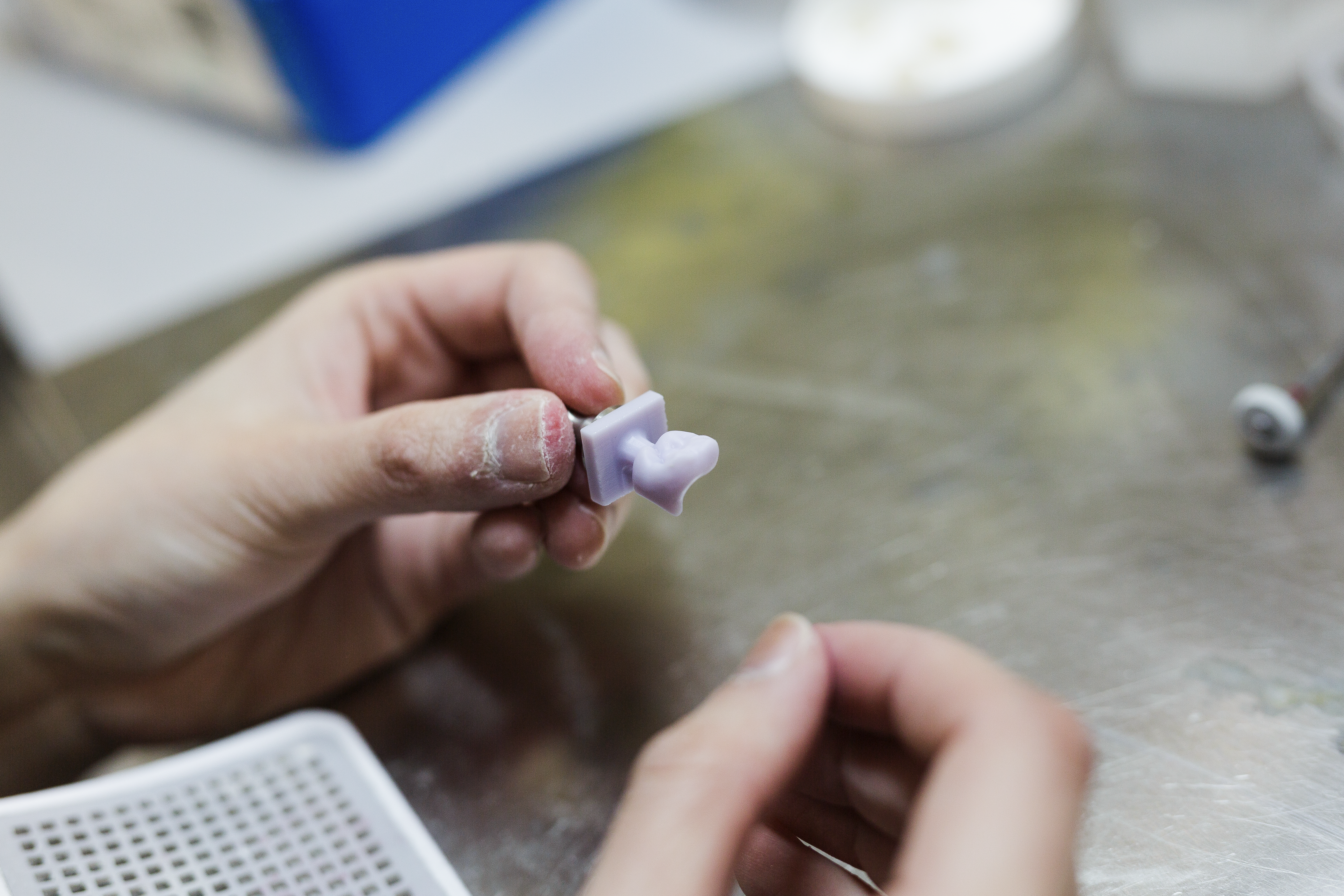 A dental technician or dentist holds a small lavender-tinted dental crown or bridge shade guide sample between their fingers for color matching and selection.