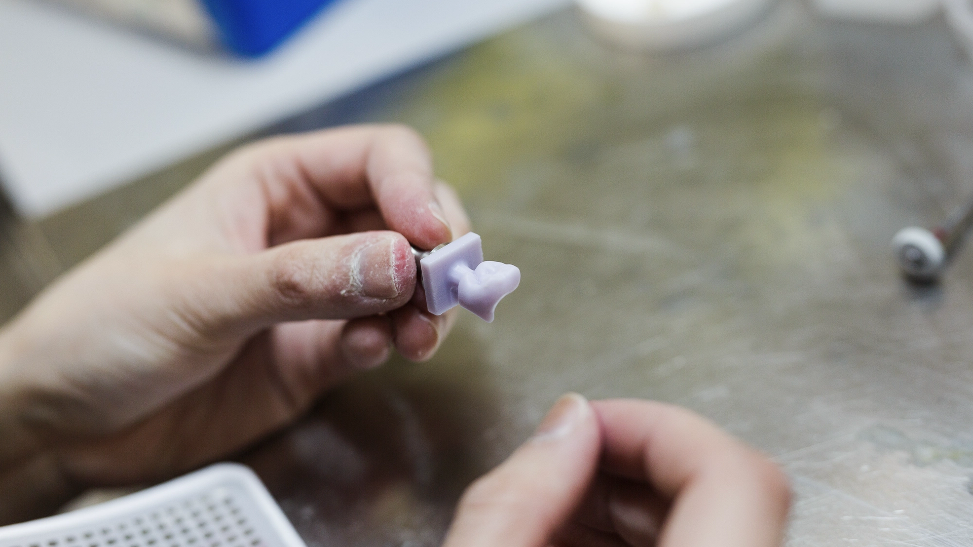 A dental technician or dentist holds a small lavender-tinted dental crown or bridge shade guide sample between their fingers for color matching and selection.