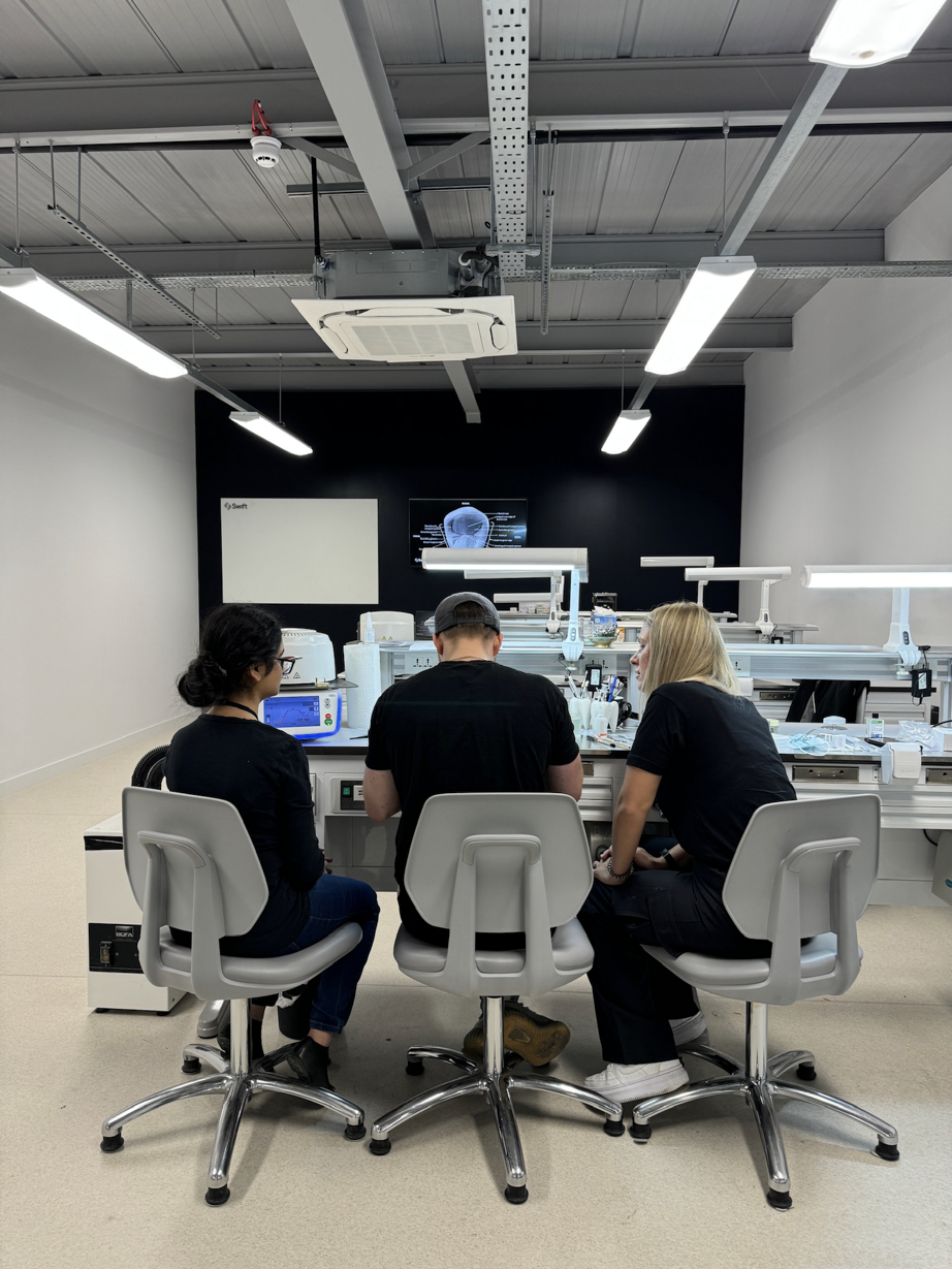Three dental laboratory technicians seated at a modern workstation with CAD/CAM milling equipment, computer displays, and dental prosthetic fabrication devices in a bright lab environment.