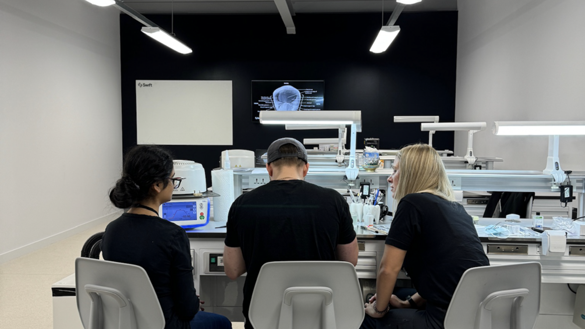 Three dental laboratory technicians seated at a modern workstation with CAD/CAM milling equipment, computer displays, and dental prosthetic fabrication devices in a bright lab environment.