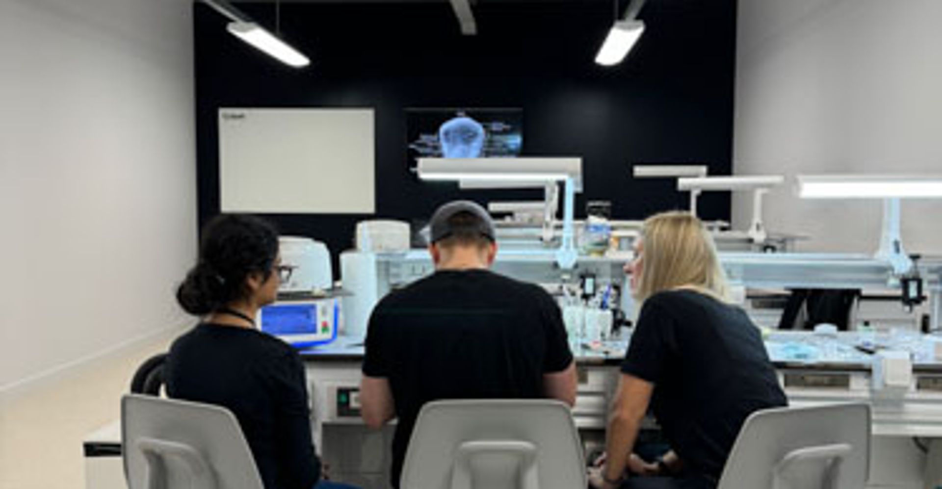Three employees sit at a desk in a contemporary office space with an industrial ceiling, fluorescent lighting, and a dark accent wall, appearing to work together on a project.