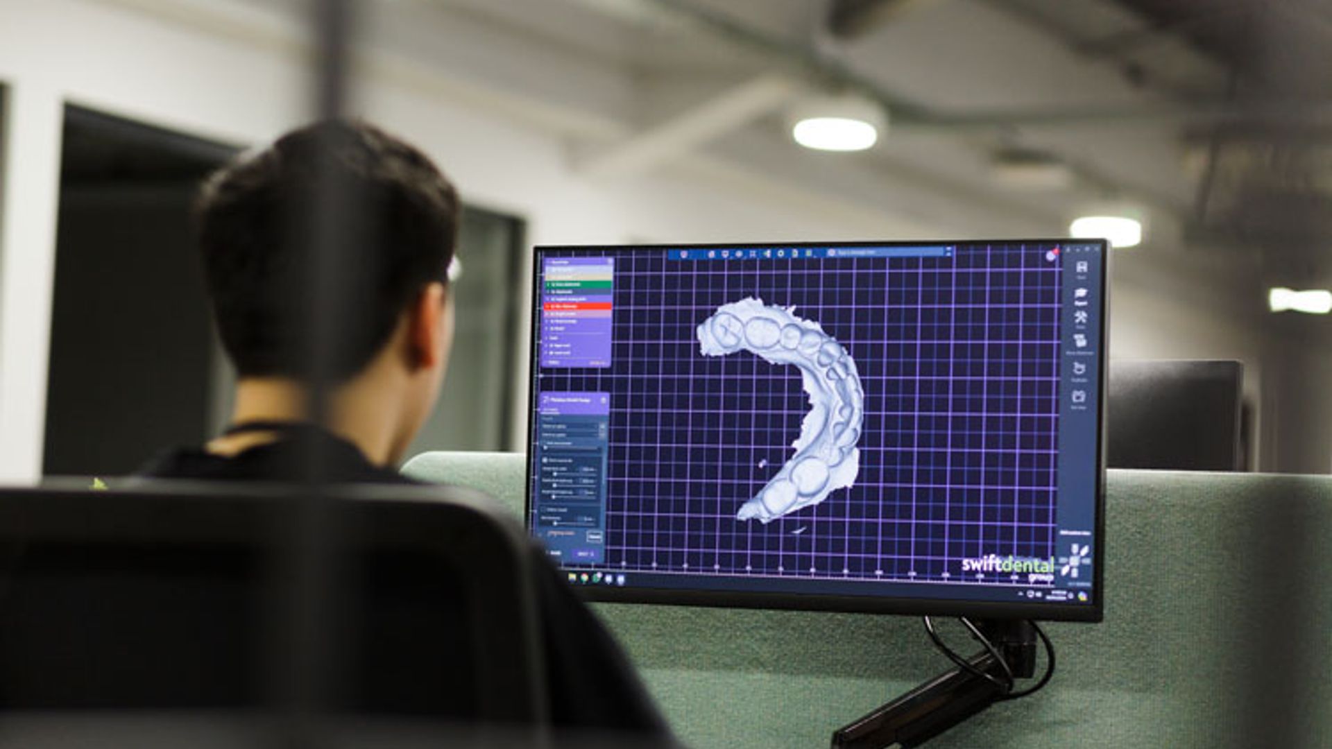 A person viewed from behind sits at a workstation viewing a computer monitor displaying a white 3D dental model on a blue grid interface in a modern office or laboratory setting.