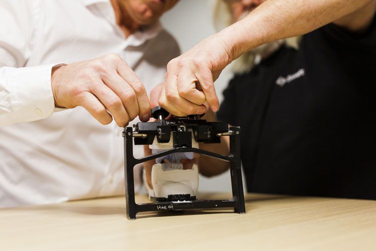 Two men in casual business attire collaborate while adjusting or demonstrating a small black 3D printer on a wooden work surface.