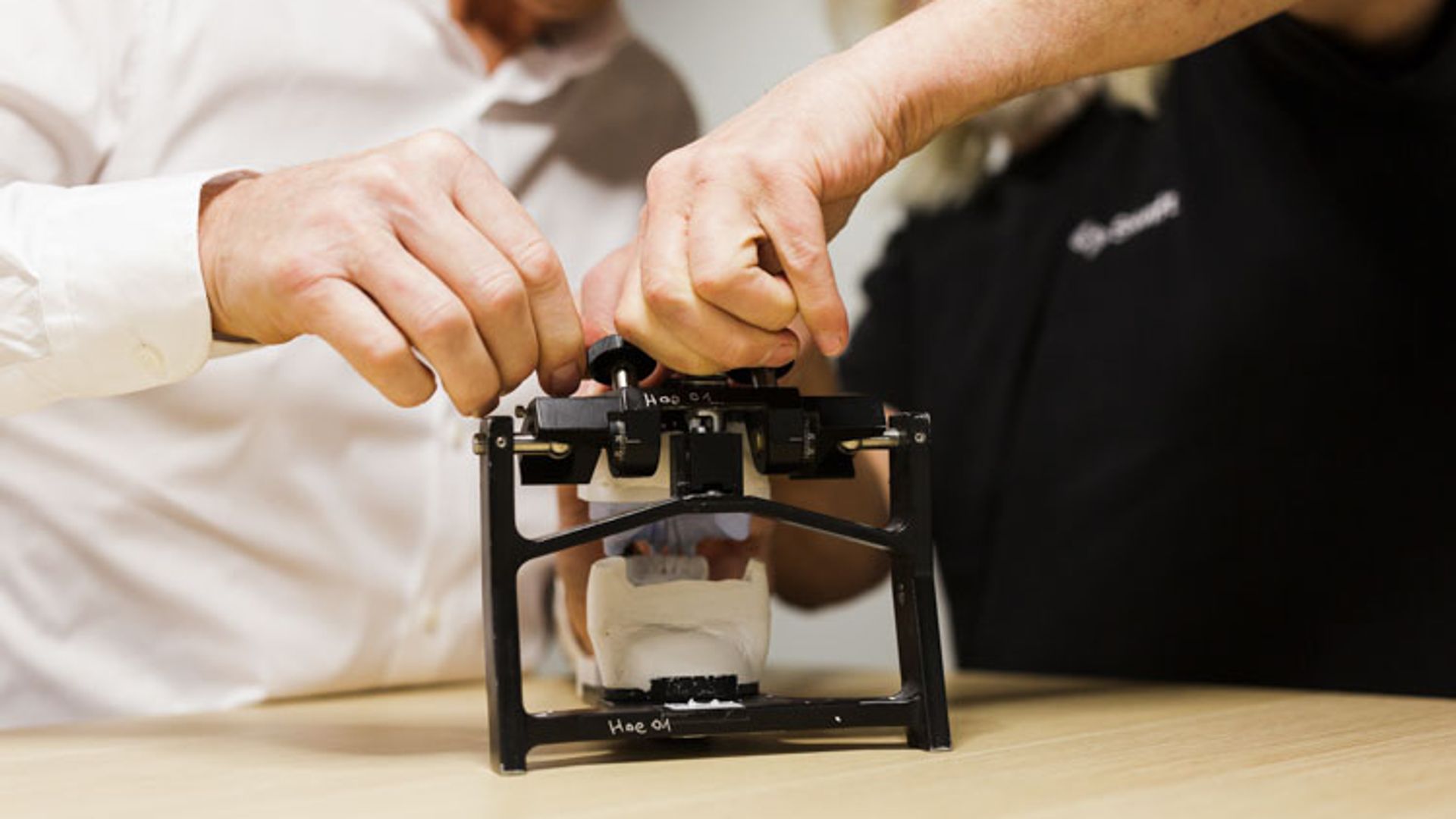 Two men in casual business attire collaborate while adjusting or demonstrating a small black 3D printer on a wooden work surface.