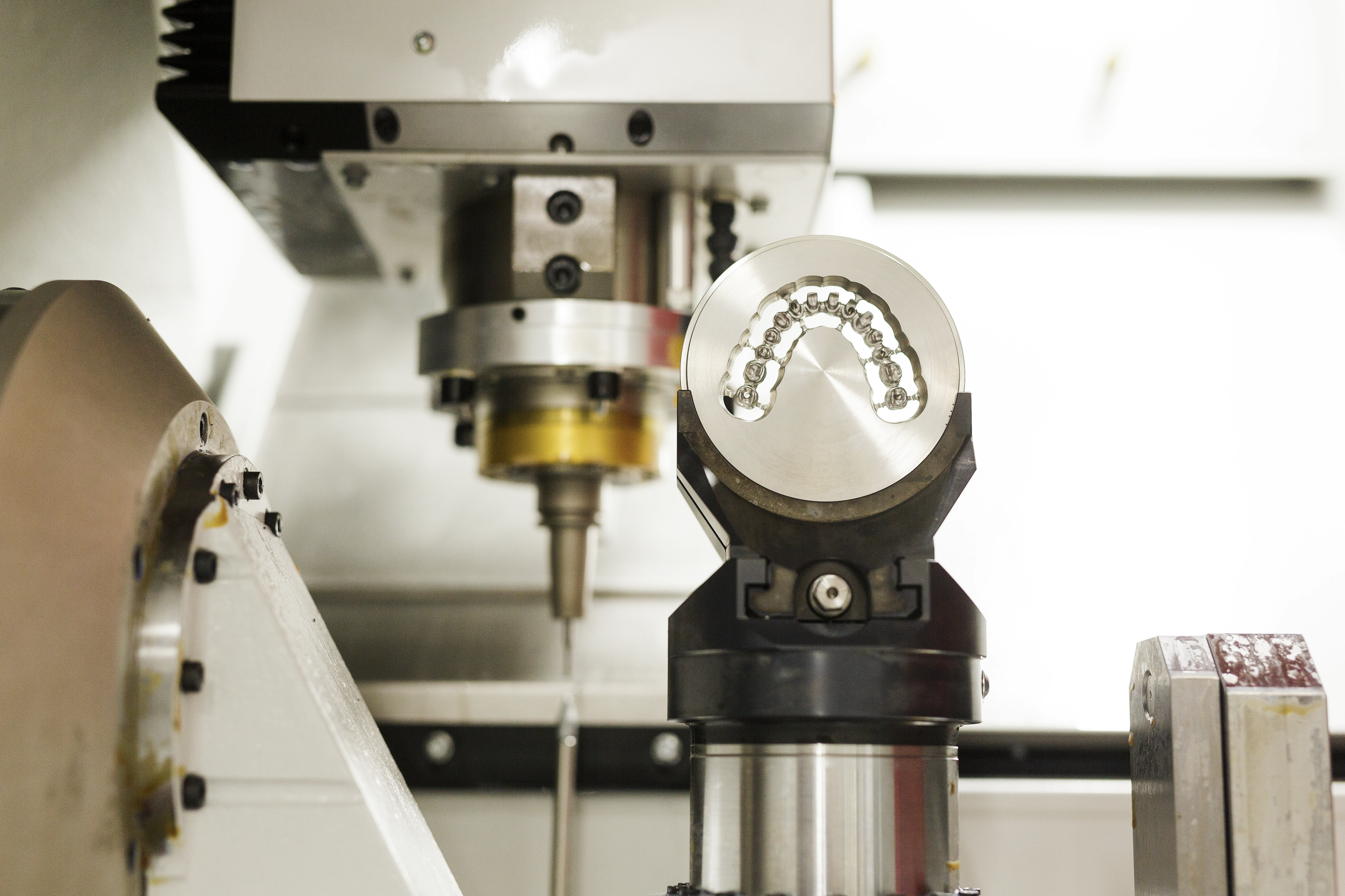 A dental CAD/CAM milling machine displays a freshly milled ceramic dental crown on its spindle, with cutting tools and rotary components visible in the background.