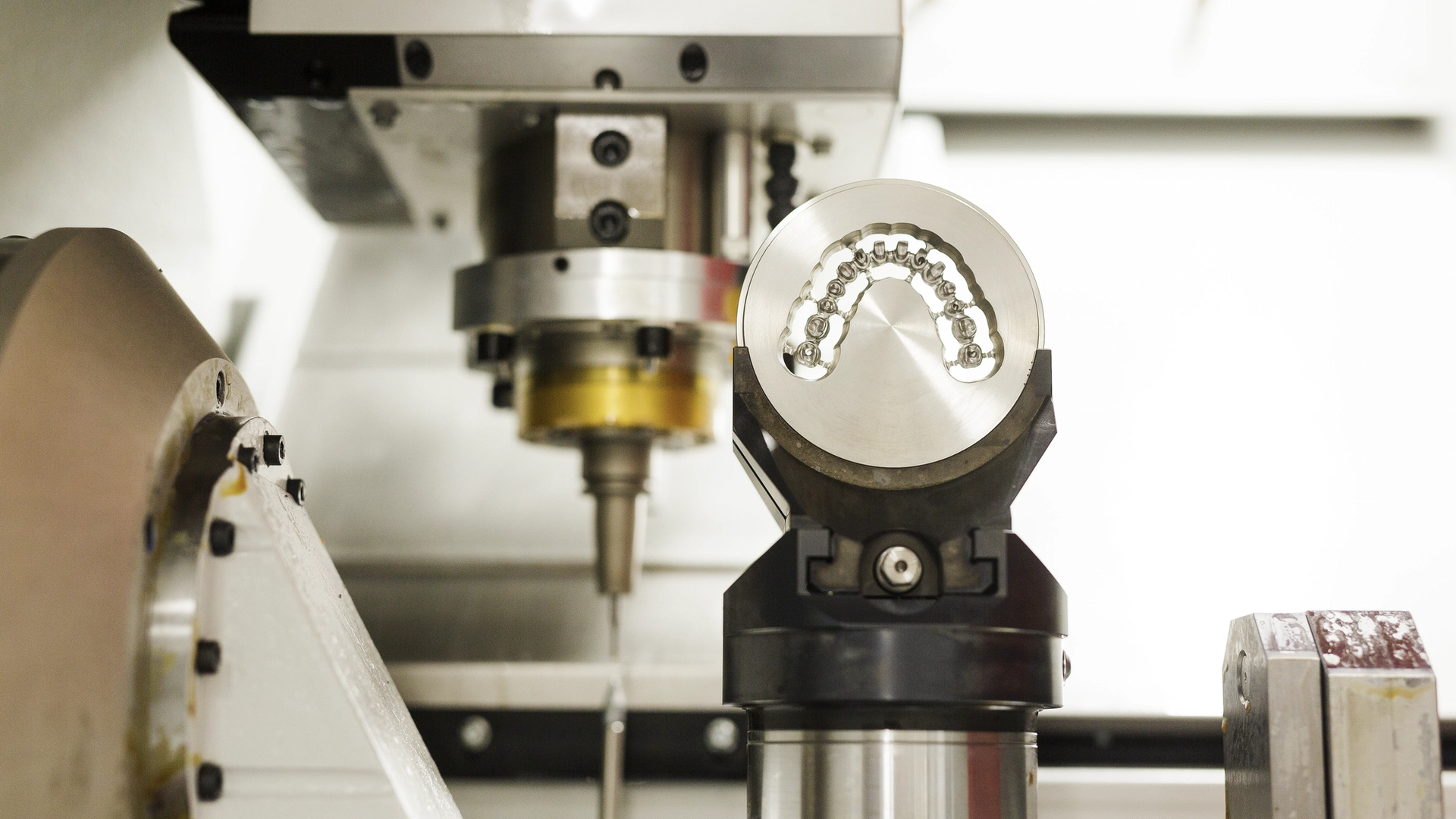 A dental CAD/CAM milling machine displays a freshly milled ceramic dental crown on its spindle, with cutting tools and rotary components visible in the background.